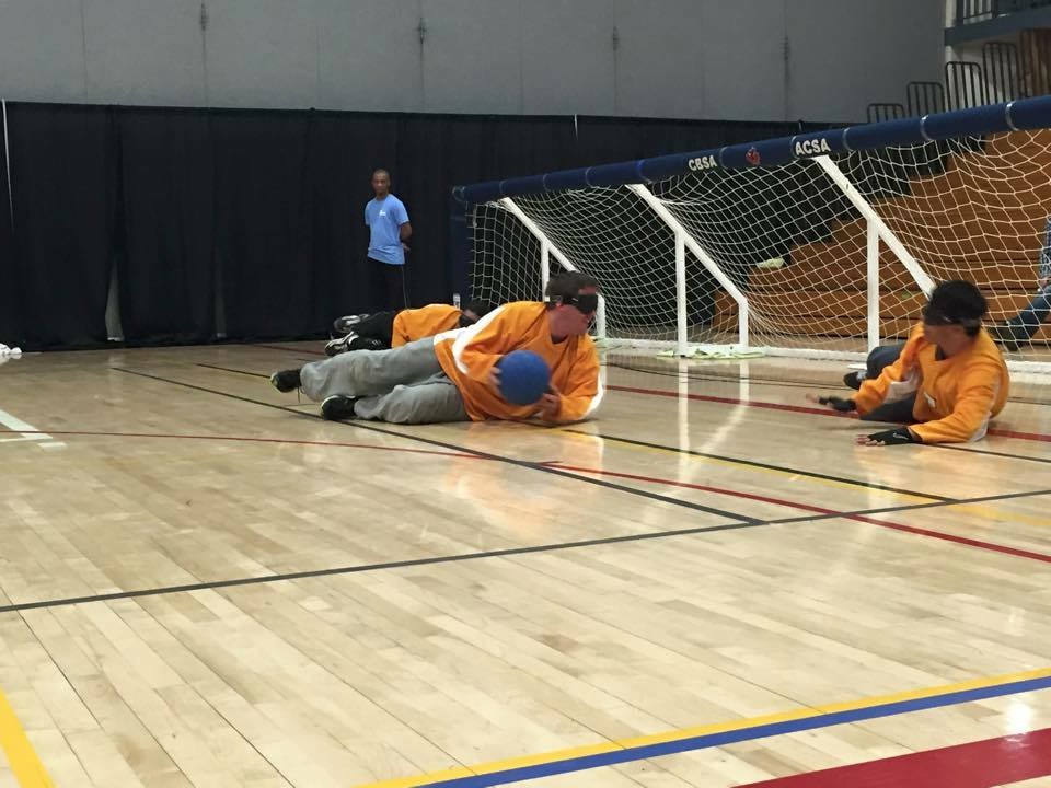 Athletes in yellow jerseys playing goalball. Three players lay on the court with a net spanning the width of the court behind them. the player in the centre is about to pass to the player on his left side.
