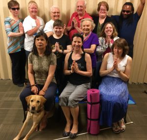 12 of our 2017-2018 season yoga participants pose for a photo, many with hands folded. A yellow lab guide dog sits to the front left, and a pink yoga mat is rolled standing in the centre.